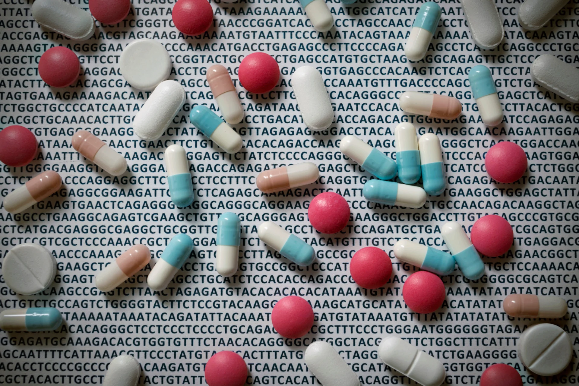 A scattering of pills over a background of repeating DNA base-pair letters, illustrating the link between genetic code and medication response.